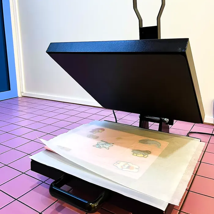 La presse à chaud de l'atelier, ouverte sur un tote bag couvert d'écussons prêt à être thermocollé